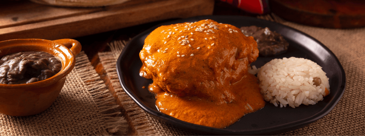 Pollo en pipián rojo con ajonjolí, acompañado de arroz blanco y frijoles negros en un plato de barro mexicano